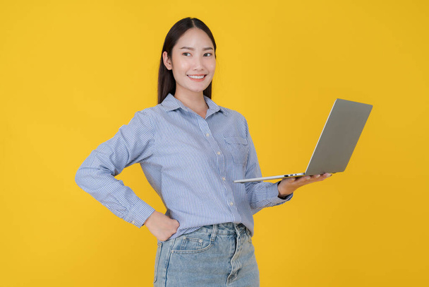 Jovem mulher asiática inteligentemente vestida com uma camisa listrada azul sorri enquanto segura um laptop cinza, de pé contra um cenário amarelo vibrante, exalando confiança e profissionalismo - Foto, Imagem