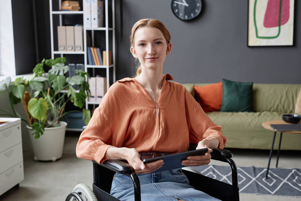 Portret van een jonge vrouw met een rolstoel die lacht naar de camera en een tablet vasthoudt, kopieerruimte - Foto, afbeelding