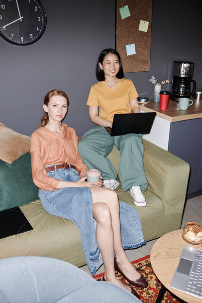 Вертикальный полноразмерный портрет двух современных молодых женщин, улыбающихся в камеру, наслаждающихся работой в офисной гостиной с вспышкой - Фото, изображение