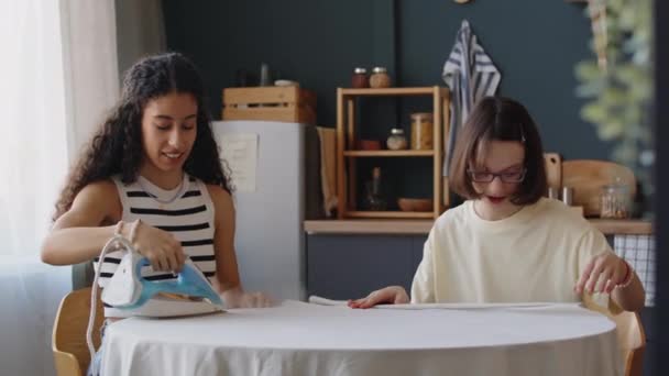 Medium shot of Caucasian girl with cerebral palsy ironing tablecloth at kitchen while doing household chores on weekend and Biracial female friend helping her - Footage, Video