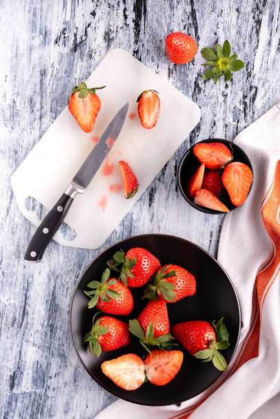 von oben, eine Schüssel und ein schwarzer Teller mit leckeren ganzen und in Scheiben geschnittenen roten Erdbeeren auf dem Schneidebrett - Foto, Bild