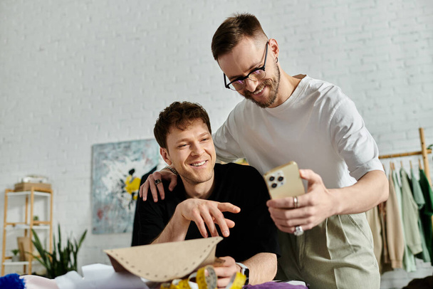 Δύο άνδρες, ένα αξιαγάπητο γκέι ζευγάρι, στέκονται περήφανα ο ένας δίπλα στον άλλο στο εργαστήρι σχεδιαστών τους, επιδεικνύοντας τις μοντέρνες δημιουργίες ενδυμασίας τους.. - Φωτογραφία, εικόνα