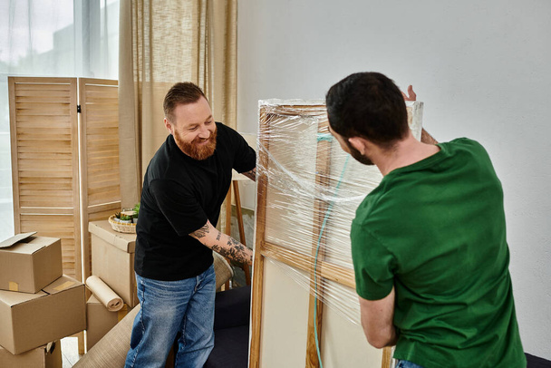 A gay couple in love standing near a frame in their new home, surrounded by moving boxes. - Photo, Image