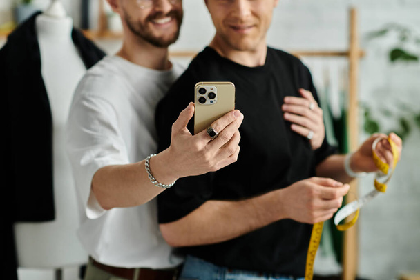 Δύο άντρες, ένα γκέι ζευγάρι, στέκονται δίπλα δίπλα με το smartphone, δουλεύοντας πάνω σε μοντέρνα ενδυμασία στο εργαστήρι σχεδιαστών τους.. - Φωτογραφία, εικόνα