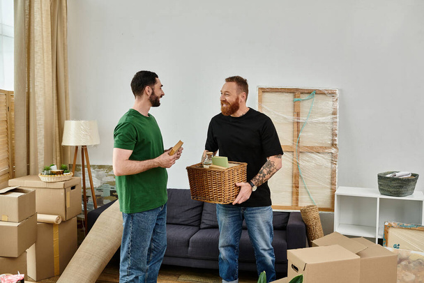 A gay couple embraces in their new living room, holding a basket, surrounded by moving boxes, beginning their new life together. - 写真・画像