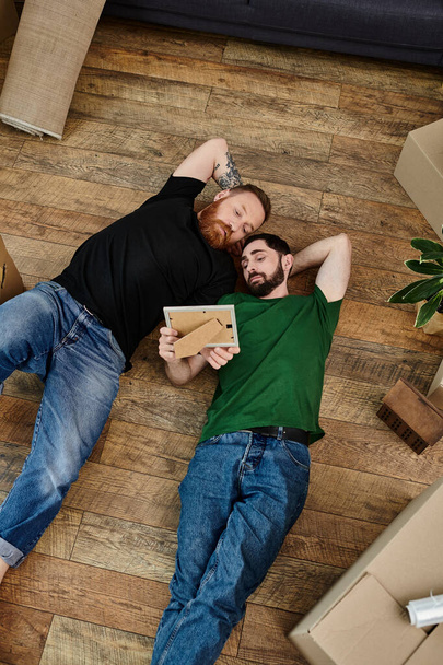 A loving gay couple laying tenderly on a wooden floor amid moving boxes in their new home. - Valokuva, kuva