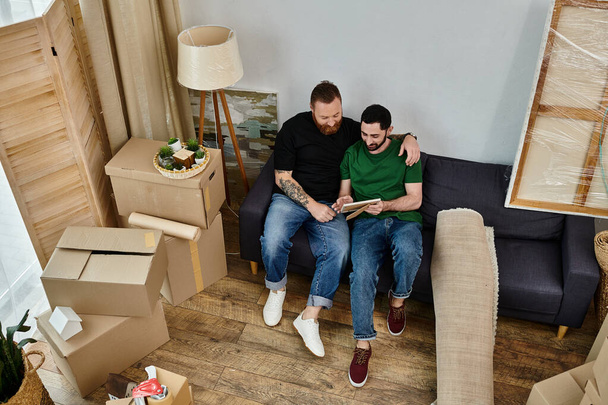Ένα γκέι ζευγάρι κάθεται στην κορυφή ενός καναπέ, αγκαλιάζοντας το νέο τους σπίτι ανάμεσα σε κουτιά, συμβολίζοντας την αγάπη και τα νέα ξεκινήματα.. - Φωτογραφία, εικόνα