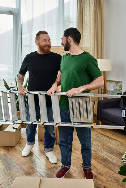 Δύο άντρες, ένα γκέι ζευγάρι, στέκονται μαζί στο σαλόνι τους και κρατούν σκάλα, ξεκινώντας ένα νέο κεφάλαιο στη ζωή τους.. - Φωτογραφία, εικόνα