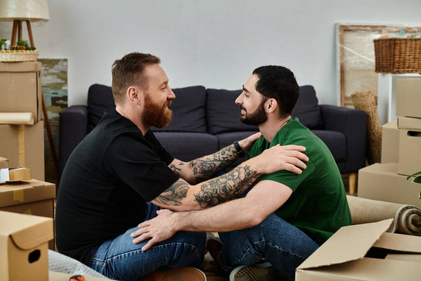 Ένα ερωτευμένο ζευγάρι γκέι κάθεται μαζί σε στοιβαγμένα κουτιά, συμβολίζοντας ένα νέο κεφάλαιο στη ζωή τους καθώς μετακομίζουν στο νέο τους σπίτι. - Φωτογραφία, εικόνα