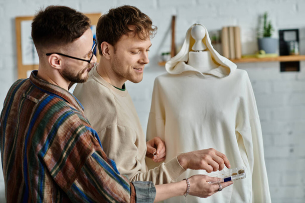 Dos hombres, una pareja gay enamorada, examinan cuidadosamente un vestido elegante en un maniquí en su taller de diseño. - Foto, Imagen