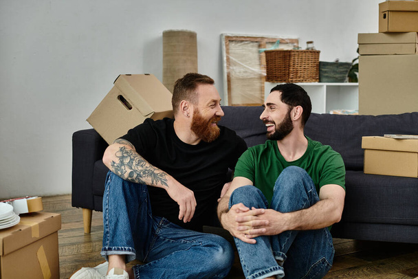 Ein schwules Paar, verliebt, sitzt auf einer Couch, umgeben von Umzugskartons, und beginnt ein neues Leben zusammen. - Foto, Bild