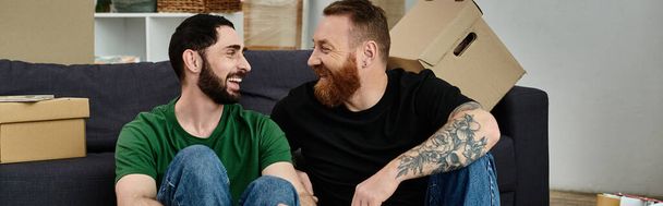 A gay couple lovingly embraces while sitting on top of a cozy couch in their new home surrounded by moving boxes. - Фото, изображение