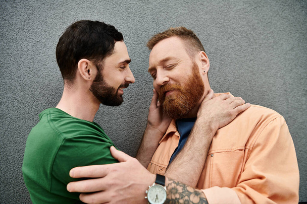 Two men, dressed casually, stand side by side against a grey wall, displaying signs of love and affection. - Foto, afbeelding