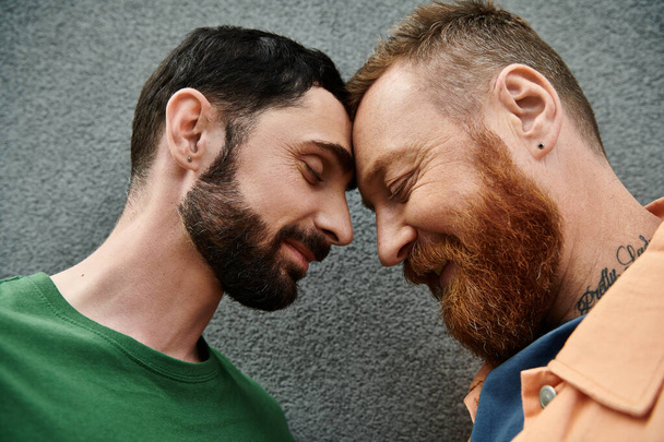 A gay couple in love, dressed in casual attire, standing closely together against a gray wall. - Photo, Image