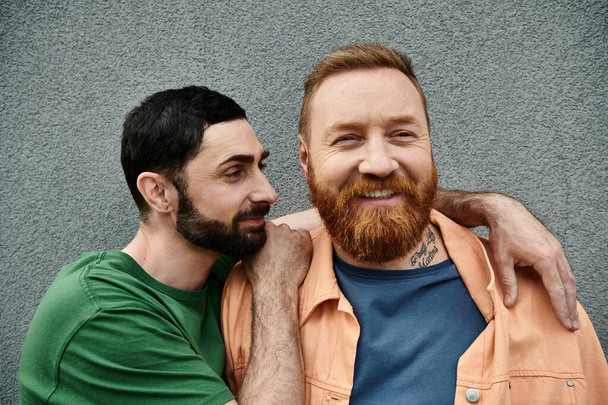 Deux hommes, un couple gay amoureux, se tiennent côte à côte en tenue décontractée, partageant une tendre étreinte contre un mur gris. - Photo, image