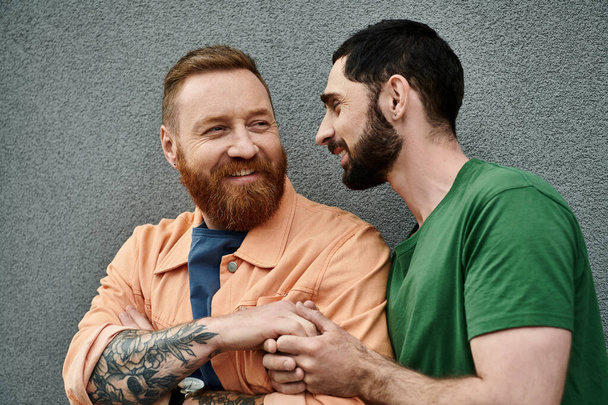 Due uomini in abbigliamento casual stanno uno accanto all'altro, mostrando affetto contro un muro grigio. - Foto, immagini