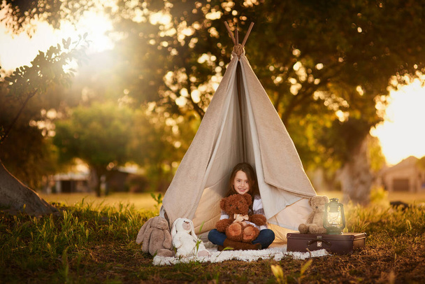 Teepee, tent and girl with toy in garden in nature for fun, playing and camping for adventure or imagination. Cloth, shelter or fantasy house for child in forest with blanket, suitcase and teddy bear. - Photo, Image