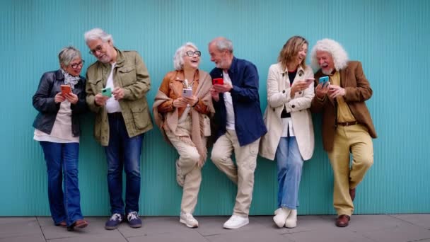 Gruppo di anziani caucasici che ridono insieme usando il cellulare appoggiato alla parete blu. Felice vecchi amici con i capelli grigi riuniti divertendosi cercando mobile all'aperto. Maturo persone godendo di dispositivi - Filmati, video