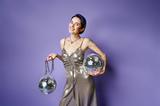 A stylish woman in a silver dress holding two disco balls, radiating glamour and fun in a studio setting. - Фото, зображення