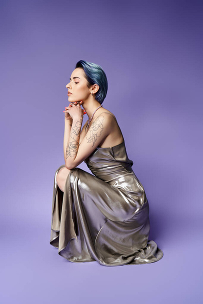 A young woman with blue hair is seated on a purple background, striking a pose in a silver party dress. - Foto, imagen