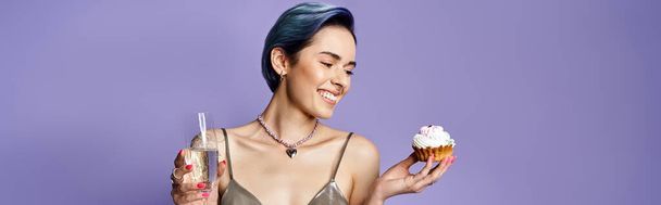 A stylish woman with short blue hair holds a cupcake and a drink in a silver party dress, exuding elegance and delight. - Fotó, kép