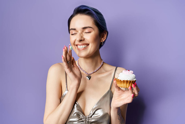 Young woman with short blue hair in a silver party dress holding a vibrant cupcake, exuding elegance and charm. - Фото, изображение