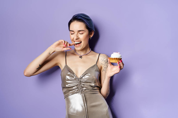 A young woman with short blue hair elegantly holds a cupcake, showcasing a silver party dress in a studio setting. - Foto, imagen