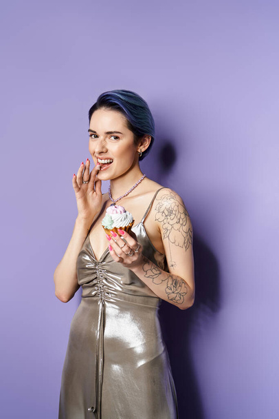 Young woman with short blue hair wearing silver dress elegantly holds a cupcake in a studio setting. - Foto, imagen