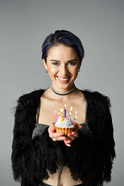 Young woman with short dyed hair in stylish attire holds up a cupcake adorned with candles, looking happy and celebratory. - Fotó, kép