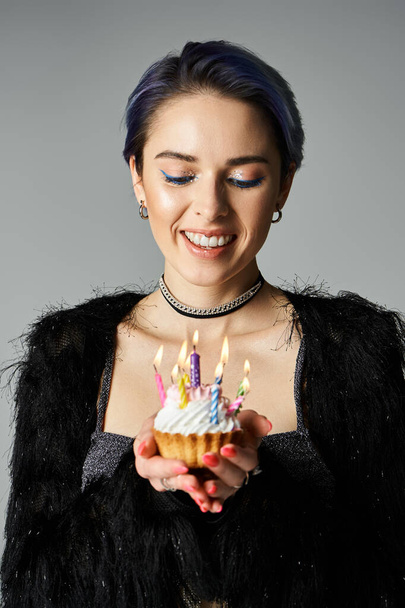 A young woman with short dyed hair holding a festive cupcake with lit candles, dressed elegantly in a studio setting. - Fotoğraf, Görsel