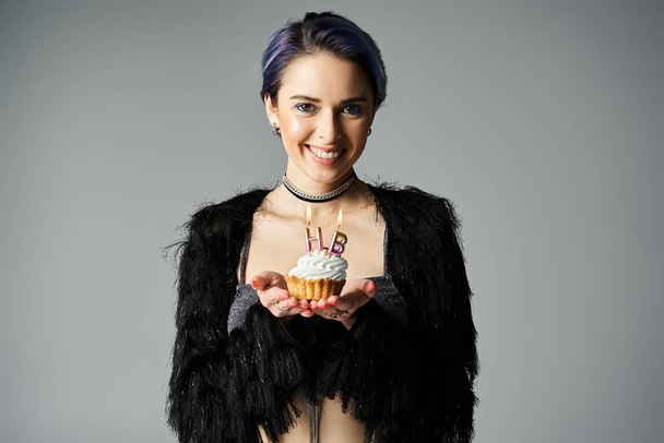 Young woman with short dyed hair holding a cupcake with icing, in stylish attire, in a studio setting. - Valokuva, kuva