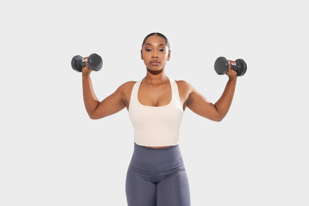 Mujer afroamericana se muestra en la imagen sosteniendo dos mancuernas en sus manos. Ella está en un entorno de gimnasio, demostrando fuerza y aptitud mediante la participación en ejercicios de levantamiento de pesas. - Foto, imagen
