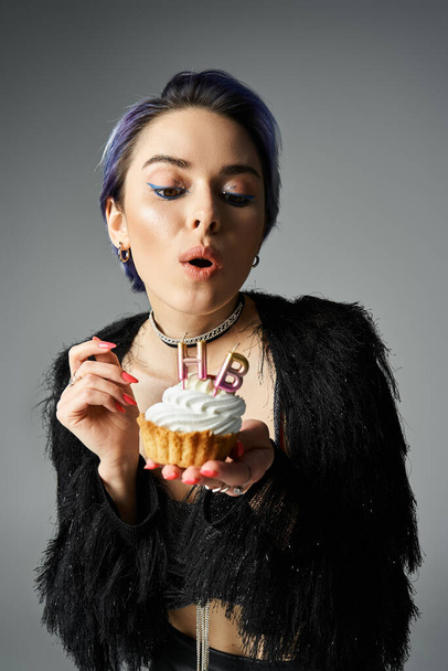 Young woman in fashionable attire holding a cupcake with a lit candle, showcasing a magical moment. - Фото, изображение