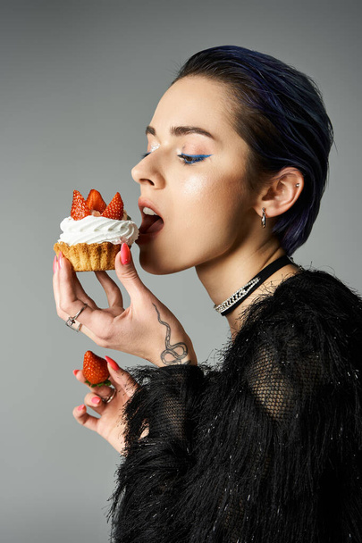 A fashionable young woman with short dyed hair enjoys a cupcake topped with strawberries in a studio setting. - Foto, imagen