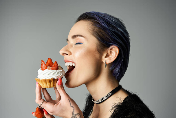 A young woman with short dyed hair smiling while eating a cupcake topped with strawberries. - Valokuva, kuva