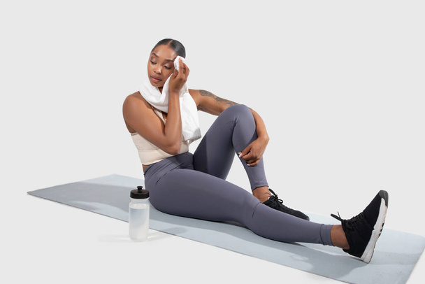 Молодая афроамериканка, одетая в тренажерный костюм, сидит на коврике для йоги с одной вытянутой ногой и опирается локтем на колено другой ноги, используя полотенце. - Фото, изображение