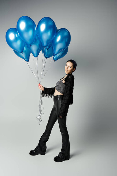 A stylishly dressed young woman with short dyed hair poses gracefully, holding a bunch of blue balloons. - 写真・画像