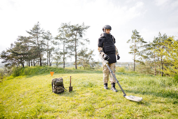 Un uomo in uniforme militare e giubbotto antiproiettile lavora nella foresta con un metal detector. Un dragamine esegue lavori di sminamento del territorio. - Foto, immagini
