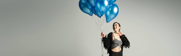 A woman with short dyed hair looks festive while holding a bunch of blue balloons in a studio setting. - 写真・画像