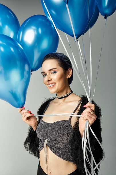 A stylish young woman with short dyed hair delightfully embraces a bunch of blue balloons in a studio setting. - Foto, Imagen