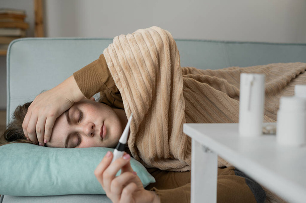 重度の頭痛と発熱を伴う病気の若い白人女性は,温度計で温度を測定し,家でソファーに横たわっています. ウイルス期間中の健康管理. - 写真・画像