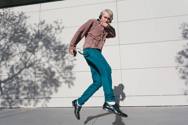 Młody Latynos z krótkimi blond włosami ubrany w retro ubrania, spodnie i brązową koszulę, na świeżym powietrzu skacze ze słuchawkami słuchając muzyki, przestrzeń kopiowania. - Zdjęcie, obraz