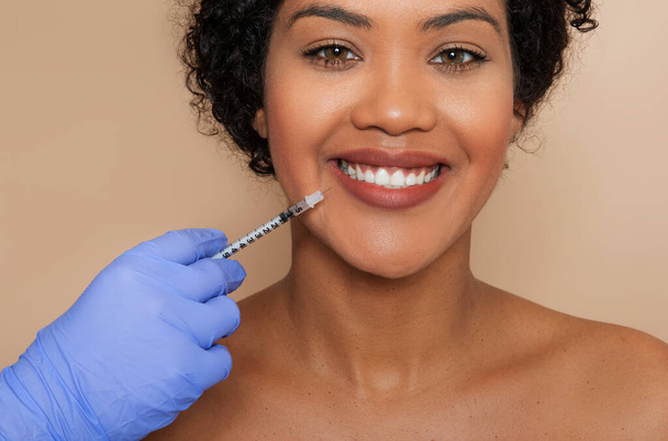 Una giovane donna sorride dolcemente mentre un operatore sanitario indossa guanti blu somministra un'iniezione cosmetica vicino alla bocca, suggerendo un trattamento dermatologico o estetico.. - Foto, immagini