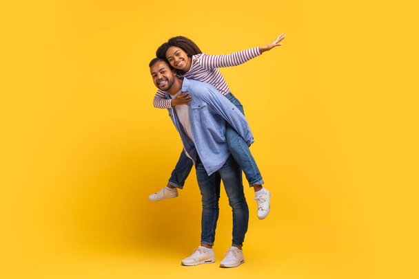 Un hombre afroamericano feliz le da a una mujer alegre un paseo a cuestas mientras ambos sonríen ampliamente, expresando disfrute despreocupado y afecto juguetón - Foto, Imagen