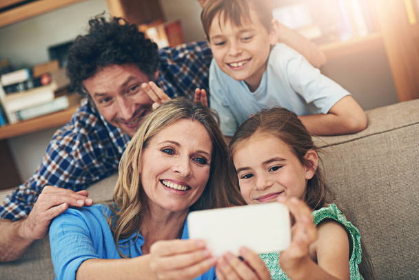 Famiglia felice, relax e selfie con segno di pace per ricordare, fotografare o legare insieme sul divano di casa. Mamma, papà e bambini piccoli con un sorriso per fotografare, catturare o momento sul divano del salotto. - Foto, immagini
