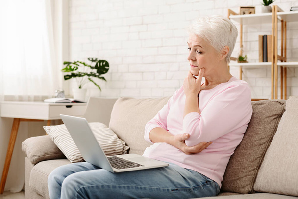 A mulher sênior está sentada em um sofá, usando atentamente um computador portátil em uma postura relaxada. Ela aparece focada na tela, com uma expressão confortável. - Foto, Imagem