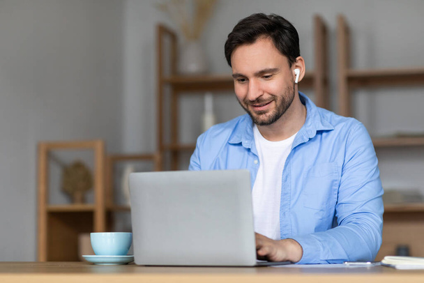 Ein Mann sitzt an einem Tisch und arbeitet mit Ohrhörern an einem Laptop. Er konzentriert sich auf den Bildschirm, tippt und blättert. Der Raum ist einfach und funktional, mit ein paar verstreuten Gegenständen. - Foto, Bild