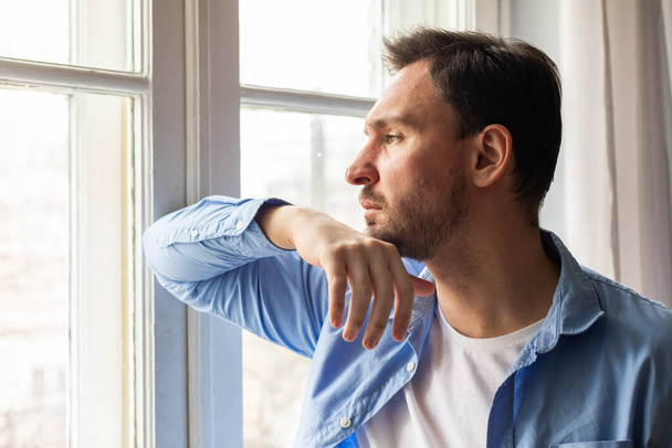 A man rests his chin on his hand as he thoughtfully looks out of a window, with gentle light filtering in, illuminating his casual attire and offering a peaceful ambiance to the start of his day. - Photo, Image