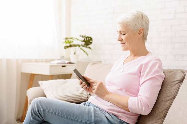 Uma mulher idosa alegre com cabelo curto está sentada confortavelmente em um sofá bege, segurando e olhando para um smartphone com uma expressão de conteúdo - Foto, Imagem
