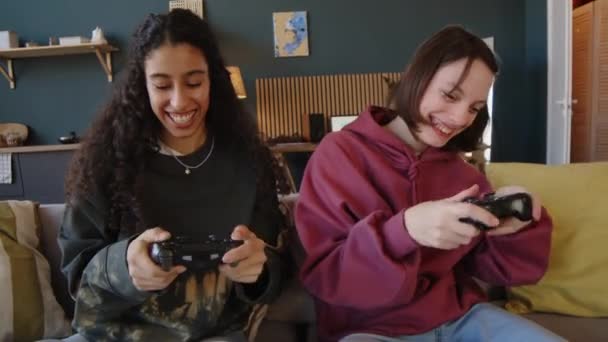 コントローラーとソファに座って一緒に楽しんでいるBiracial女性の友人と脳性麻痺のビデオゲームをしている幸せな白人十代の少女のミディアムショット - 映像、動画